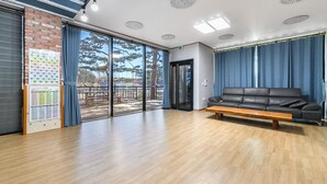 A spacious room with a wooden floor, a black leather sofa, a wooden bench, and a large window with a view of trees. - Goseong Pinetree Pension (Goseong)