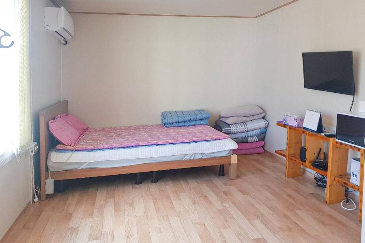 Quarto com uma cama de solteiro, uma mesa com computador e uma televisão na parede.