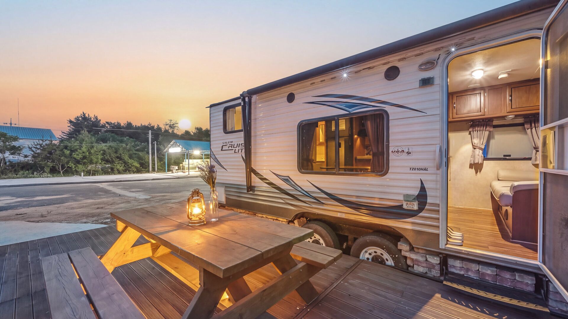 A recreational vehicle with a kitchen area, a dining table with a lantern, and a view of a building in the background.
