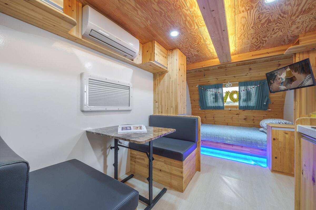 Quarto compacto com teto de madeira, cama com faixa de luz LED azul, uma mesinha com revista e televisão na parede.