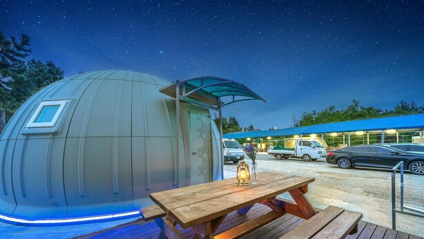 A dome-shaped structure with a transparent roof, a wooden picnic table with a lantern, and a building with a blue roof in the background. - Goseong Jjun Camping Pension (Goseong)