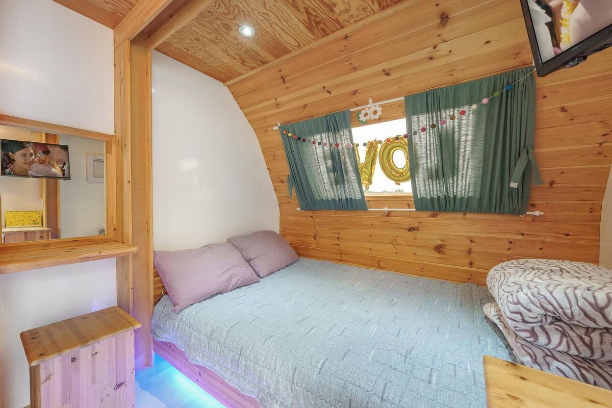 Um quarto aconchegante com teto de madeira, uma cama com cobertor cinza, um pequeno banquinho de madeira e uma janela com cortinas.