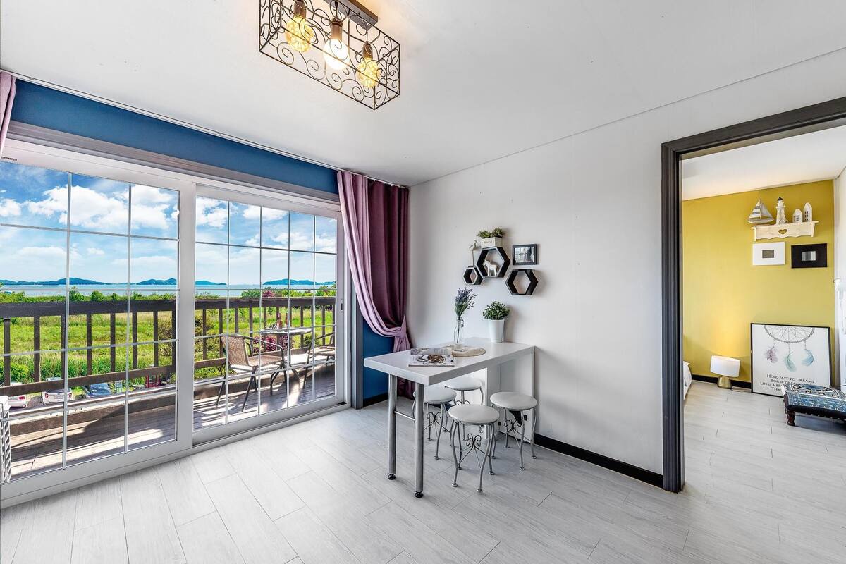 A modern room with a large sliding glass door to a balcony, a white table with bar stools, and a view of the outdoors.