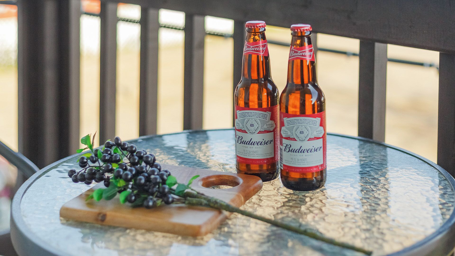 Dos botellas de cerveza Budweiser sobre una mesa de vidrio con una tabla de cortar de madera y uvas.