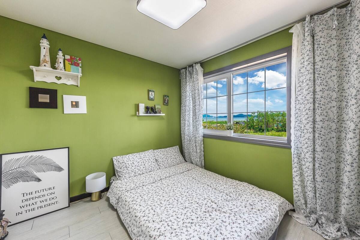 A bedroom with a bed, a window with a view, a shelf with decorative items, and a framed poster on the wall.