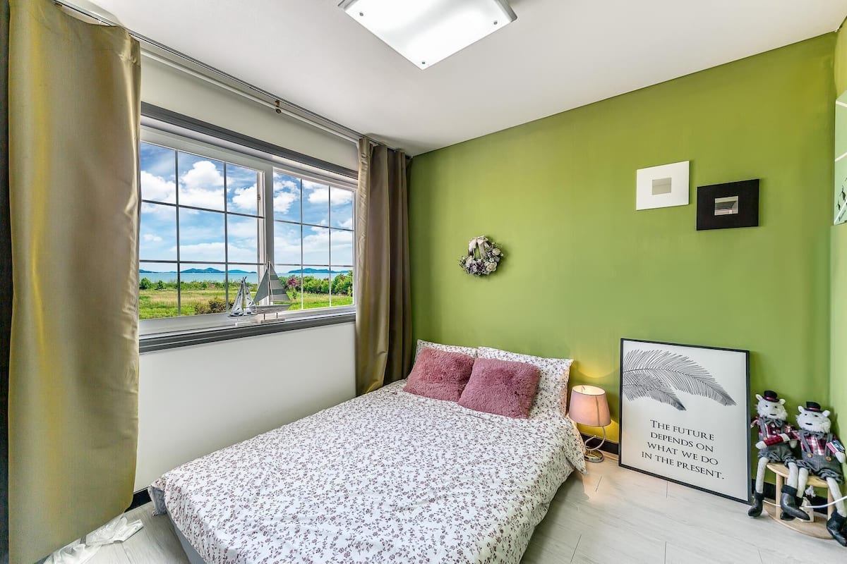 A bedroom with a bed, a window with curtains, a green wall, a framed quote, and a small shelf with figurines.