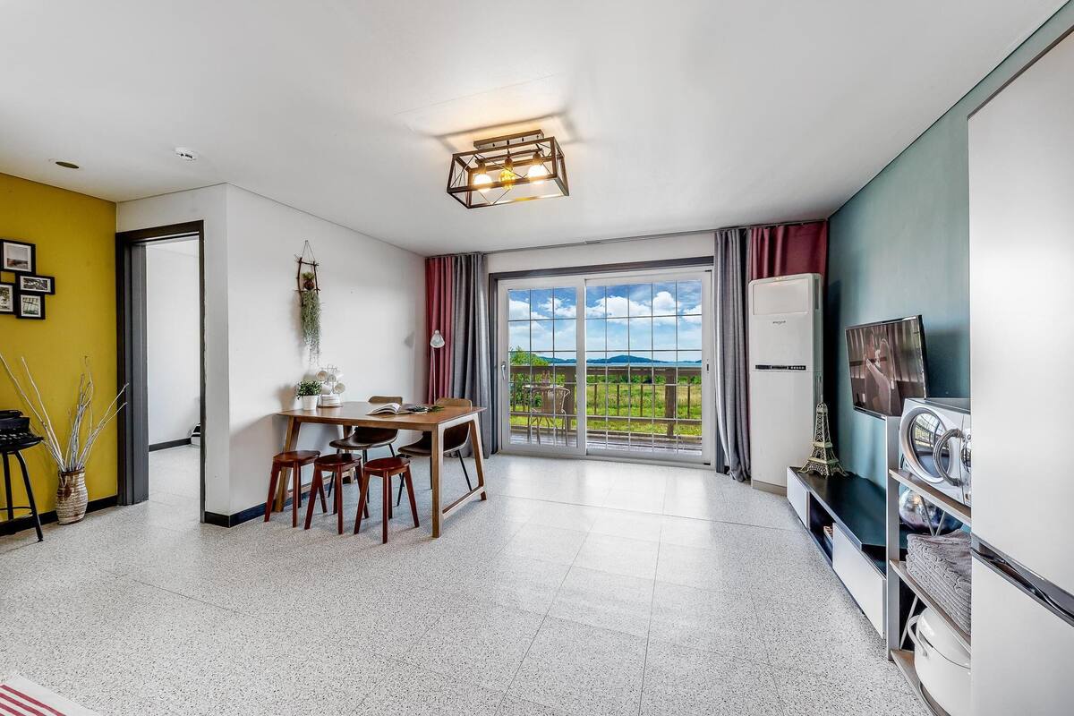 A modern living room with a dining area, a large sliding glass door leading to a balcony, and a kitchen area with a refrigerator and microwave.