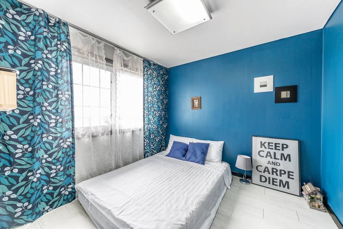 A bedroom with a bed, a window with sheer curtains, a blue wall, and a motivational poster.