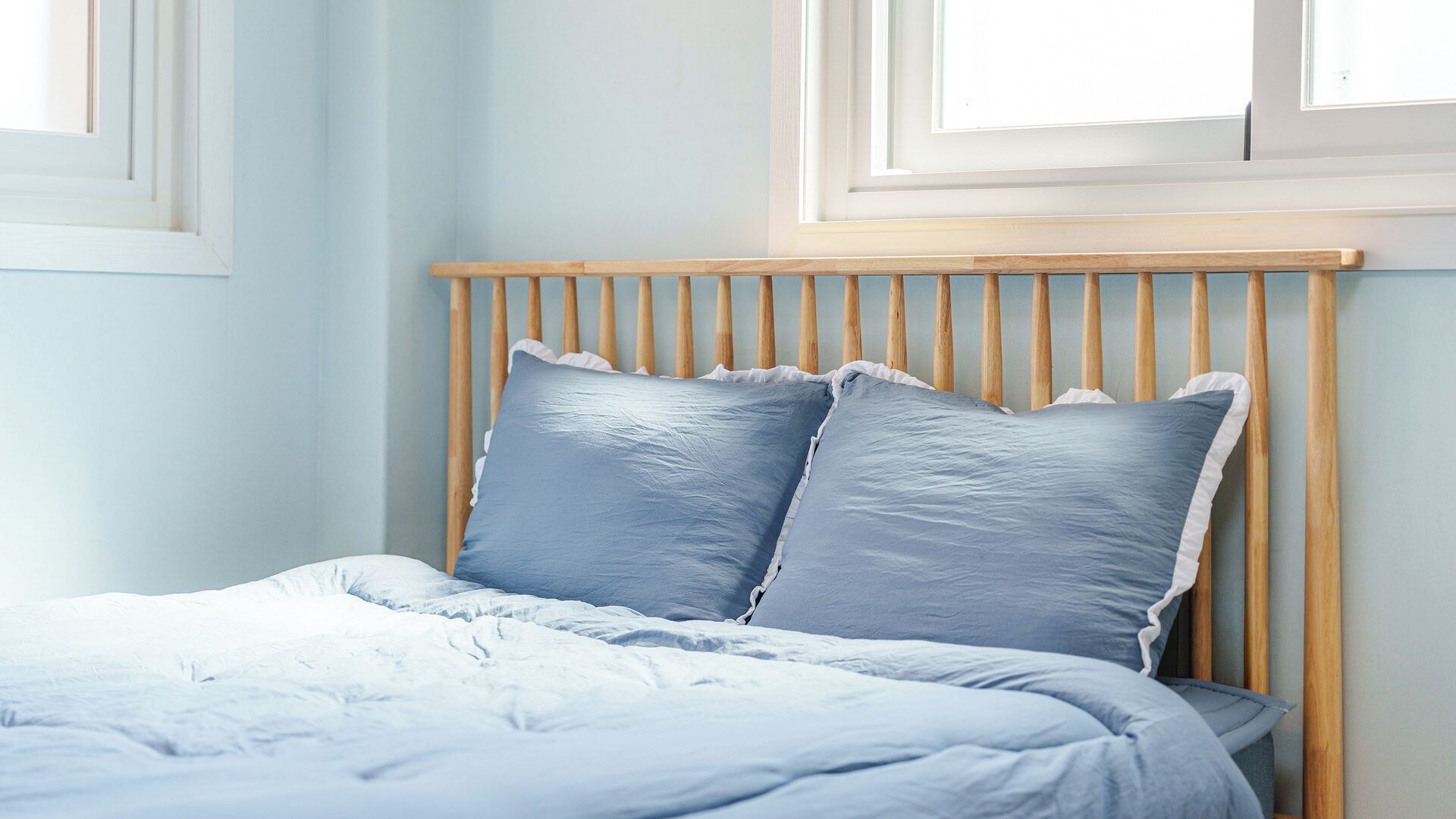Una cama con cabecera de madera y dos almohadas, frente a una pared azul claro con ventanas.