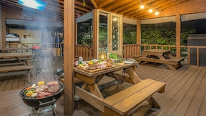 En udendørs patio med træborde og bænke, en grill med mad og udsigt til grønne områder.