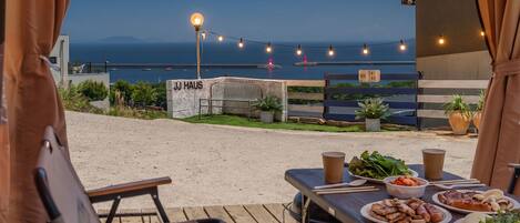 An outdoor dining area with a view of the sea, a sign reading 'JJ Haus', and string lights.