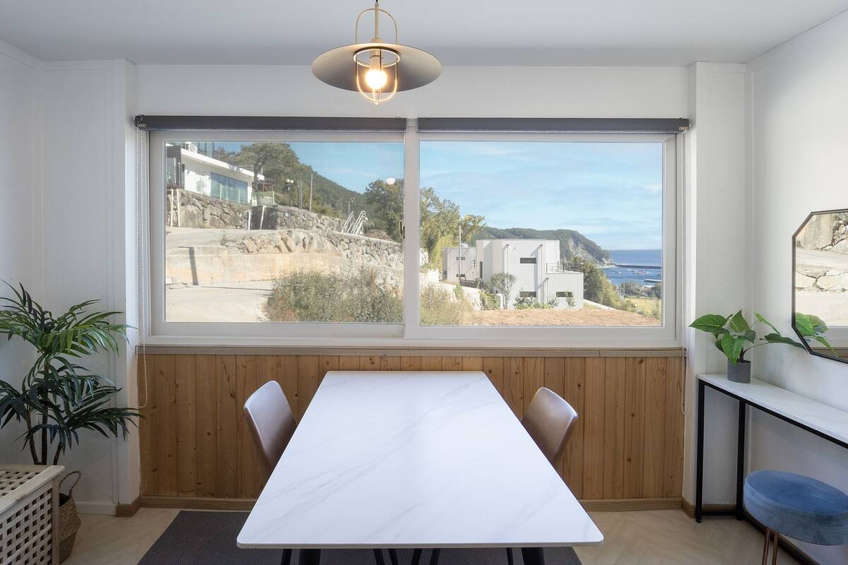 Un espace repas moderne doté d’une grande fenêtre donnant sur un paysage côtier, d’une table blanche et d’un luminaire suspendu.