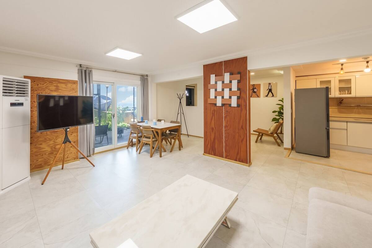 Un salon moderne doté d’un téléviseur à écran plat, d’un coin repas avec une table et des chaises, d’une cuisine équipée d’un réfrigérateur et d’un balcon aménagé avec du mobilier d’extérieur.