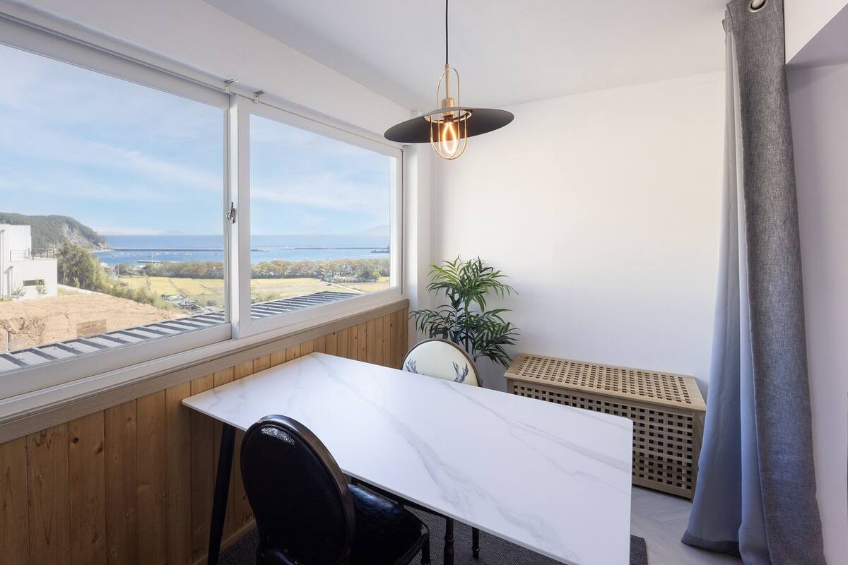 Un balcon avec vue sur la mer, une table blanche, une chaise noire et un luminaire suspendu.