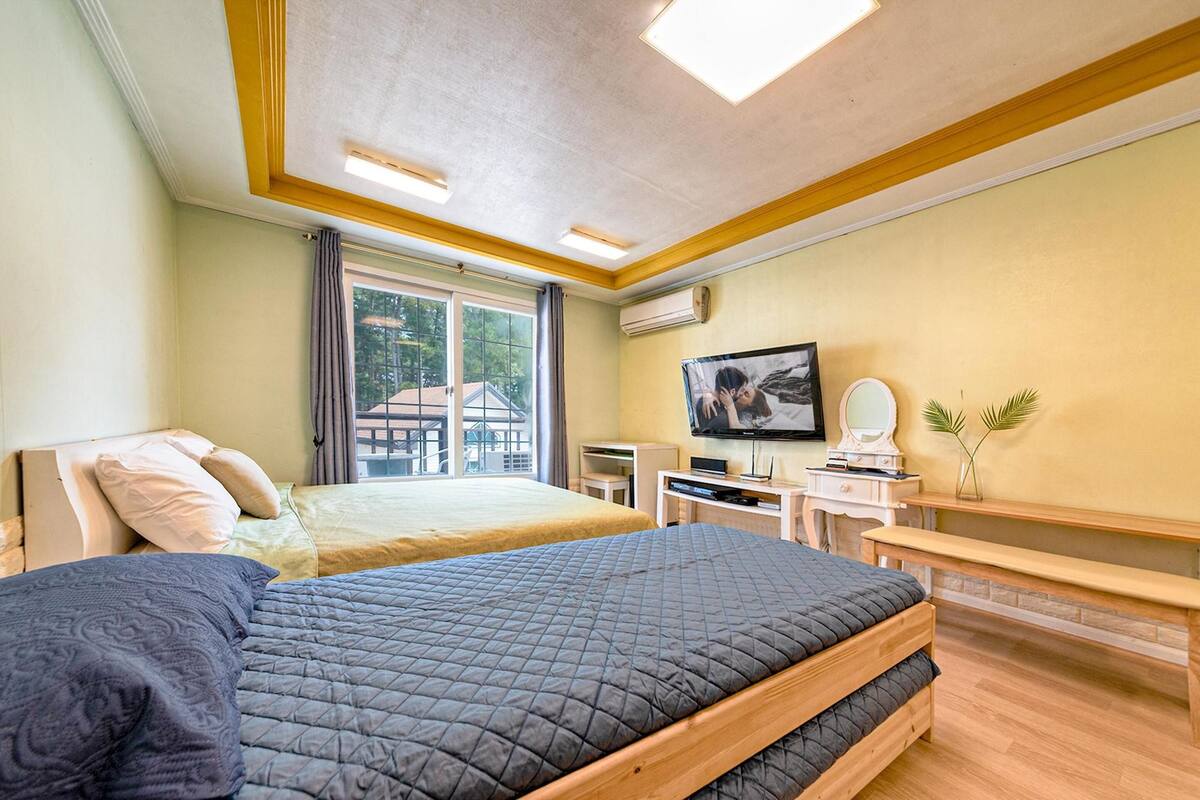 A hotel room with a large bed, a desk with a flat-screen TV, a small table, and a bench.