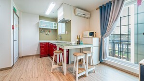 A compact kitchen with a white countertop, red cabinets, and a window with blue curtains. - Gangnueng Gadunji Pension (Gangneung)