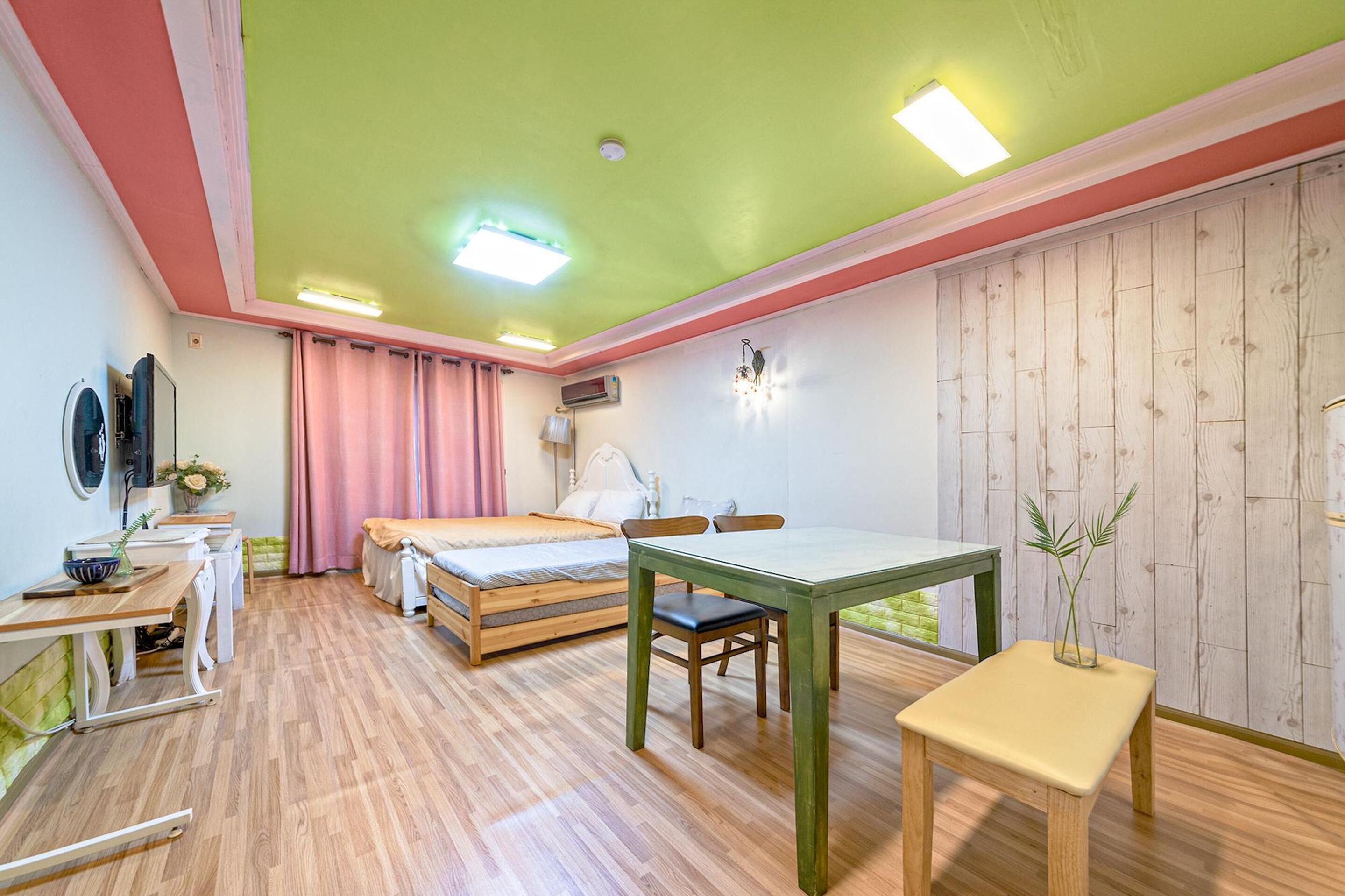 A room with a bed, a table, and a chair. There is a window with curtains, a wall-mounted air conditioner, and a small table with a vase.