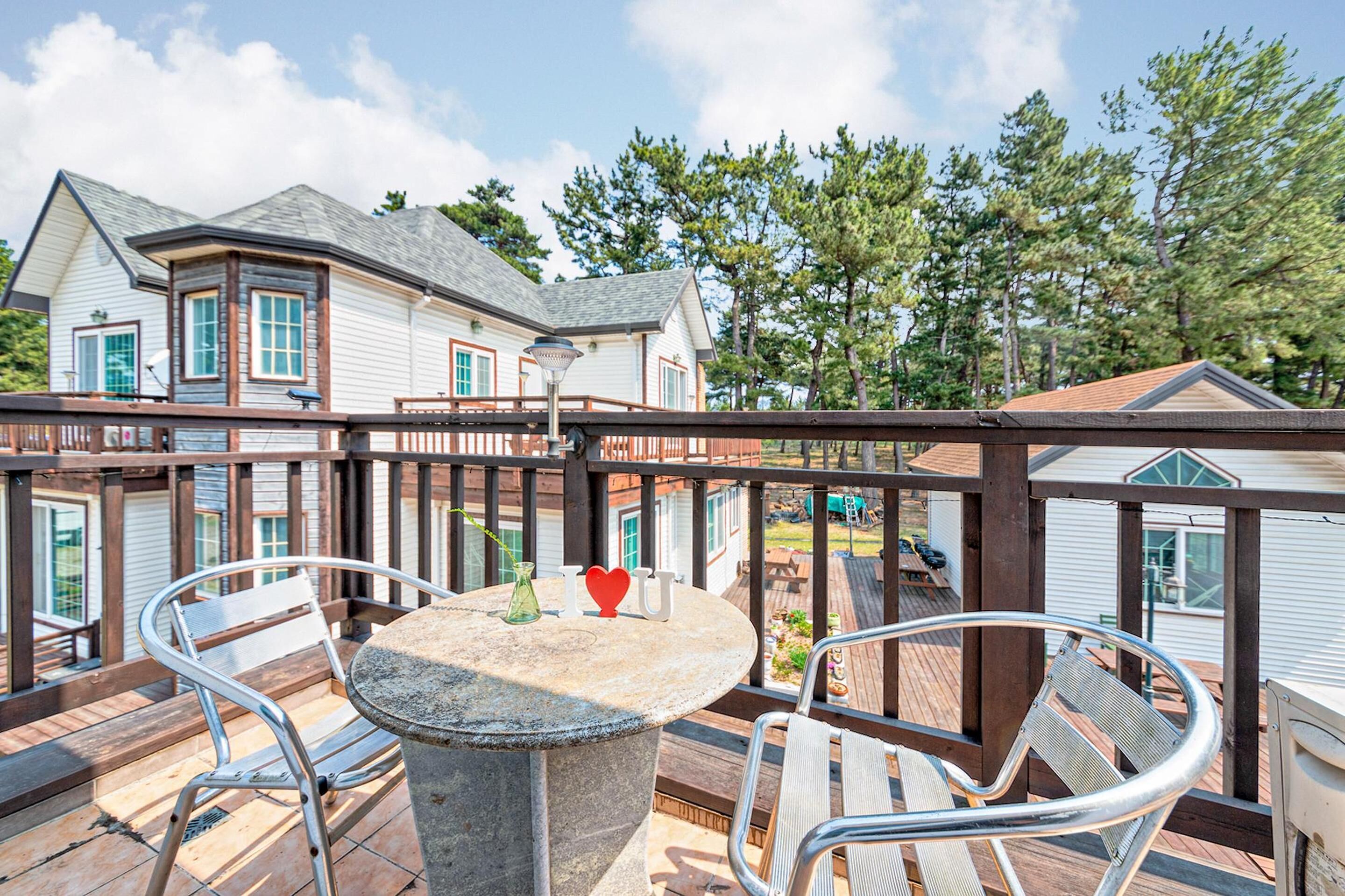Una terraza de madera con una mesa de piedra y sillas de metal, con vista a una zona residencial con casas y árboles.