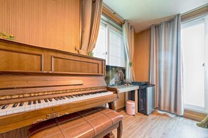 Um piano em um quarto com uma janela grande, um radiador e uma lixeira pequena.