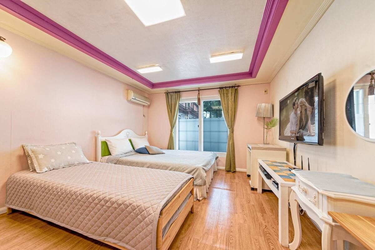 A bedroom with two beds, a desk, a TV, and a window with curtains.