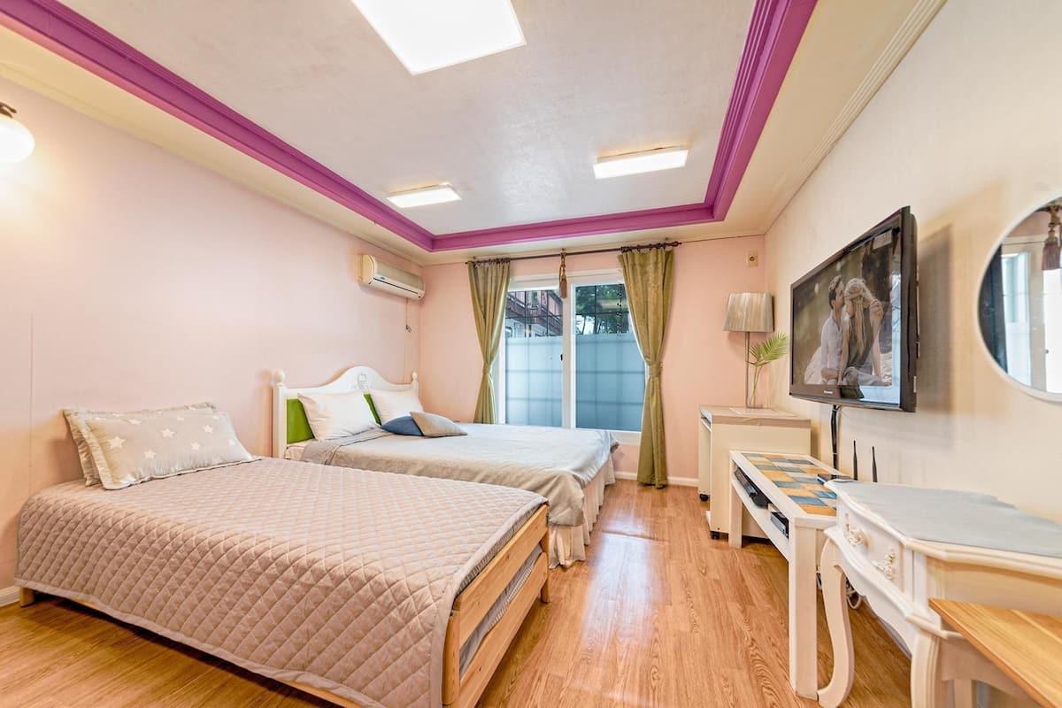 Quarto com duas camas, uma escrivaninha, uma televisão e uma janela com cortinas.