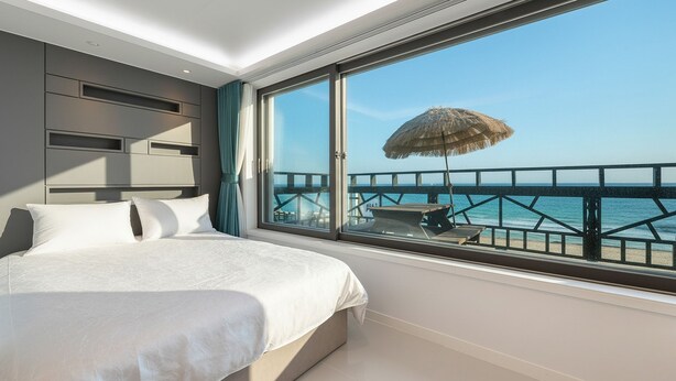 A modern hotel room with a large bed, a view of the sea, and a wooden deck with a thatched umbrella. - Gangneung Badauibyeol Pension (Gangneung)