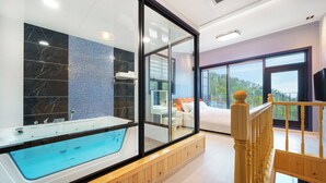 Ein modernes Badezimmer mit einer gläsernen Eckbadewanne, einem großen Spiegel und Blick auf ein Schlafzimmer mit Bett und Balkon.