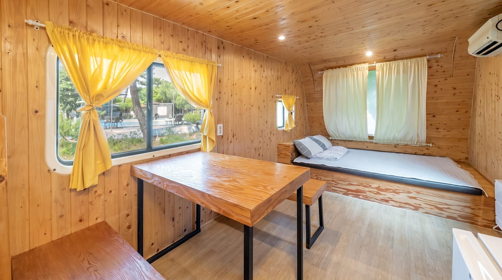 Interior de una cabaña de madera con una cama, una mesa y cortinas.