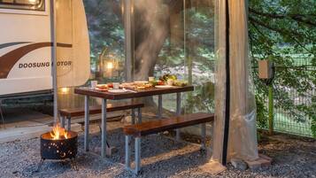 Un comedor con una mesa dispuesta para una comida, un pozo de fuego y vistas a árboles a través de un cerramiento de vidrio.