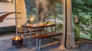 Un espace repas avec une table dressée pour un repas, un foyer extérieur et une vue sur les arbres à travers une paroi vitrée.