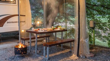 Un comedor con una mesa dispuesta para una comida, un pozo de fuego y vistas a árboles a través de un cerramiento de vidrio.