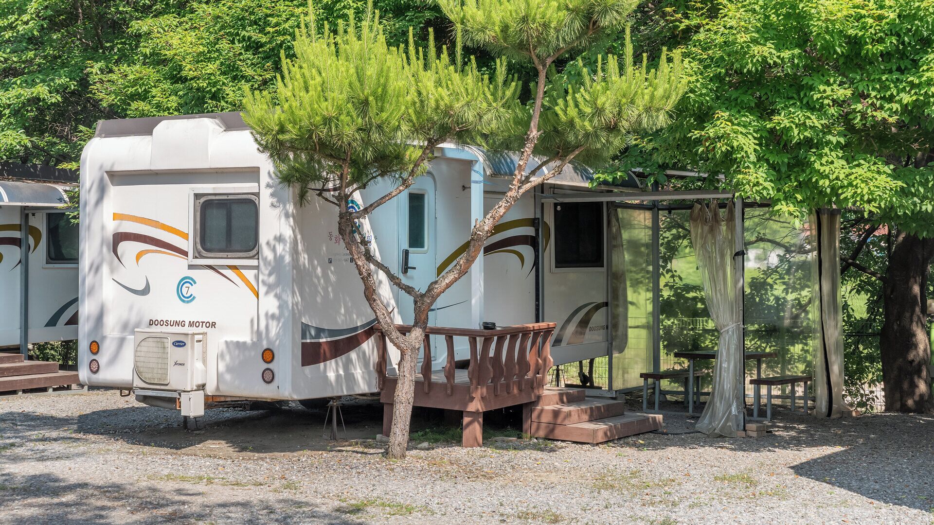 A white RV with a 'DOOSUNG MOTOR' logo, a covered porch, and a tree in front.