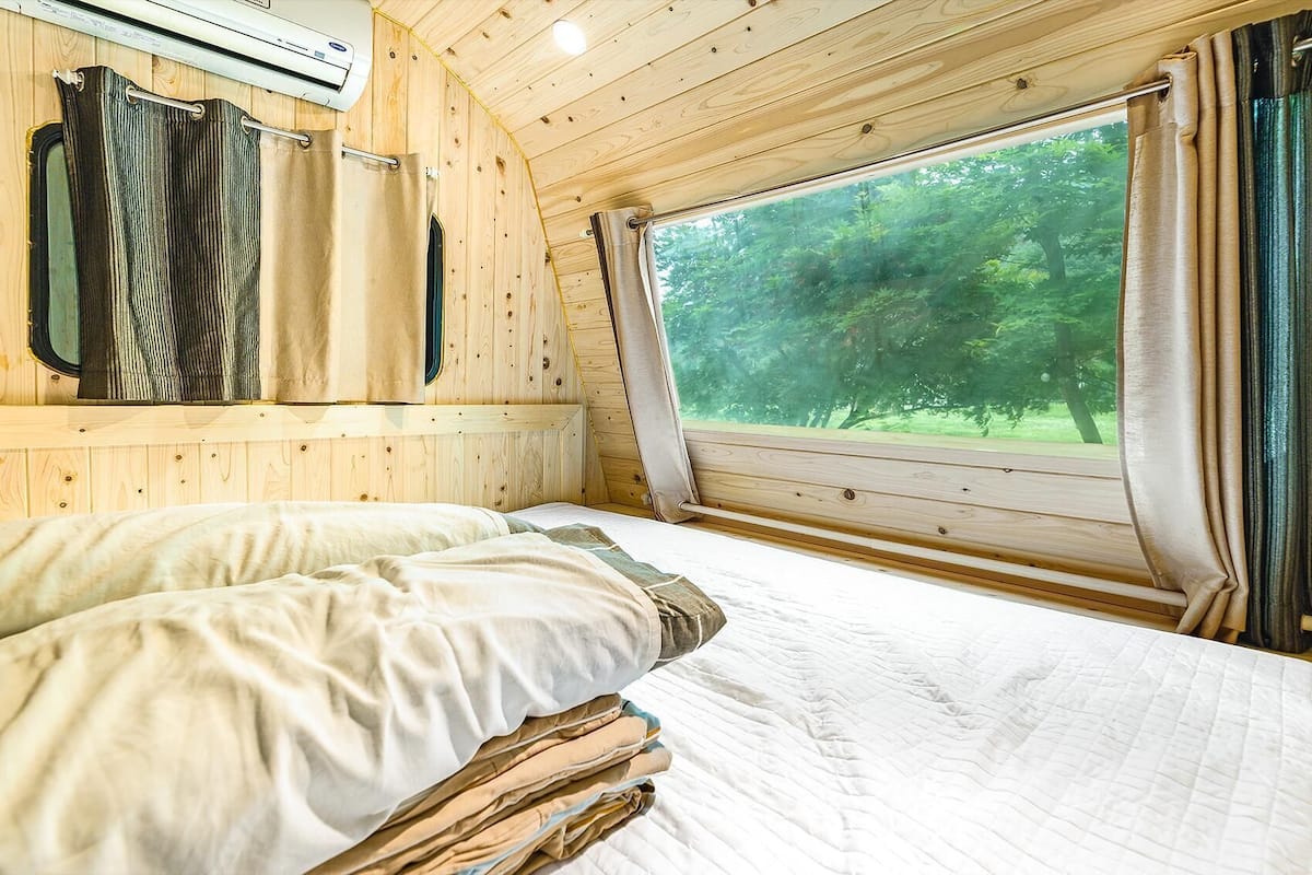 Ein Schlafzimmer einer Holzhütte mit Bett, Vorhängen und einem Fenster mit Blick auf einen Wald.