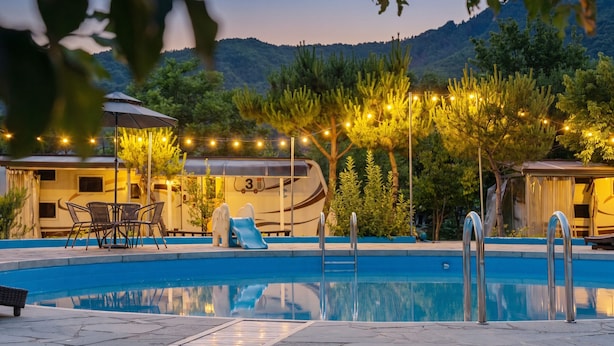 Una piscina con tobogán, rodeada de árboles y luces colgantes, con vehículos recreativos al fondo.
