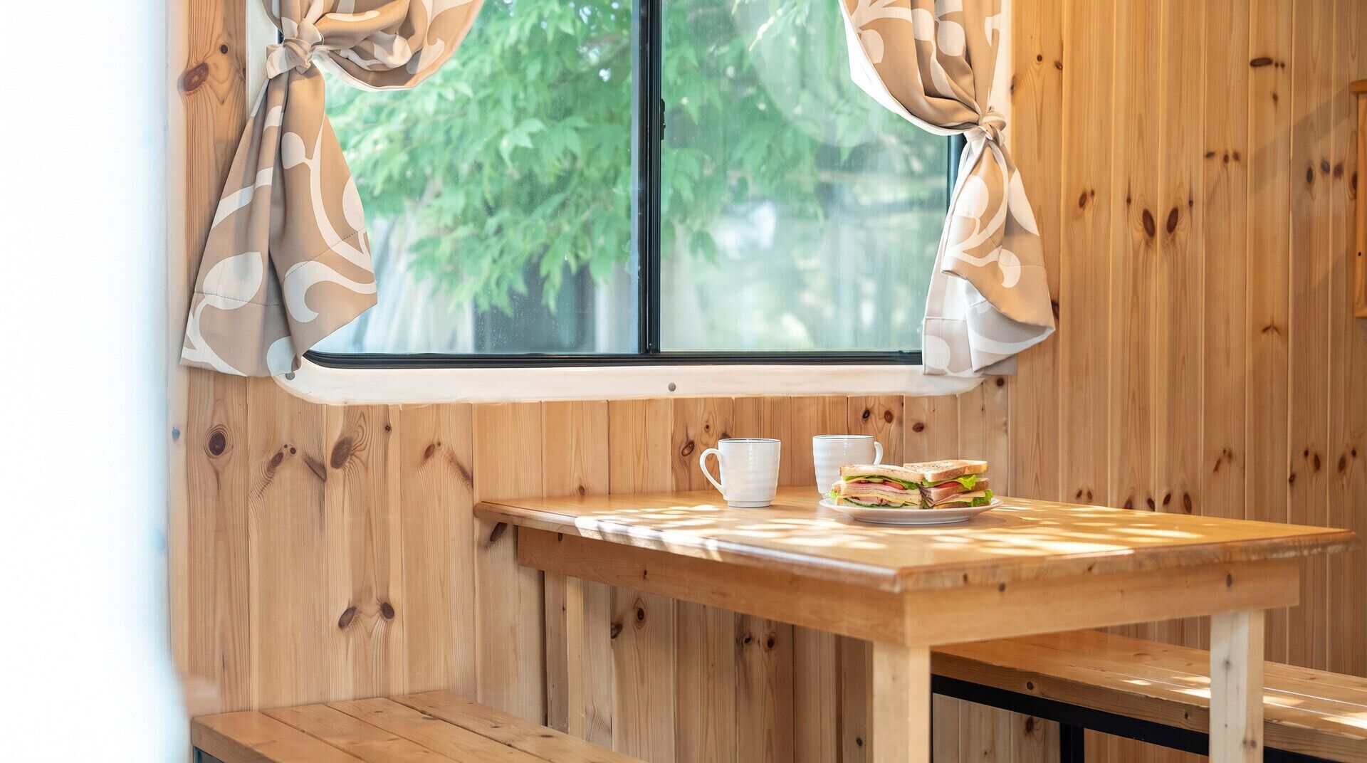 A wooden interior with a window, a table with a sandwich and two cups, and a bench.