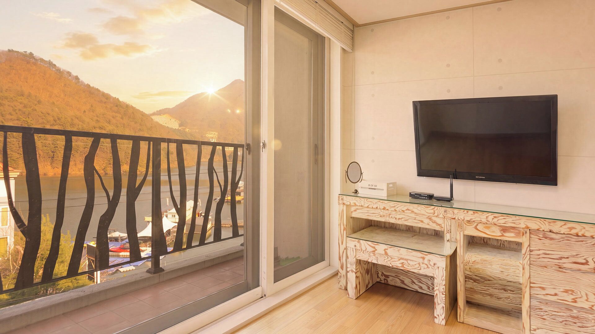 A balcony with a view of a lake and mountains, a flat-screen TV mounted on the wall, and a wooden desk with a glass top.