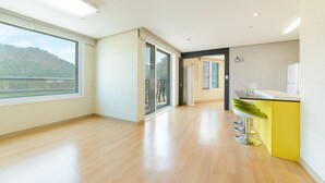 Een ruime kamer met een houten vloer, grote ramen, een schuifpui en een keukenhoek in het geel.