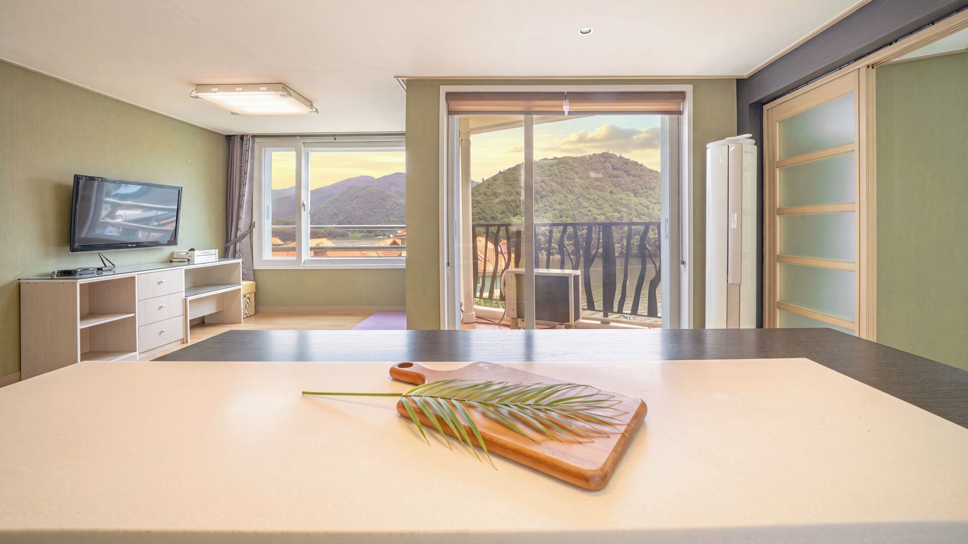 A hotel room with a large window, a flat-screen TV, a wooden table with a plant, and a sliding glass door leading to an outdoor balcony.