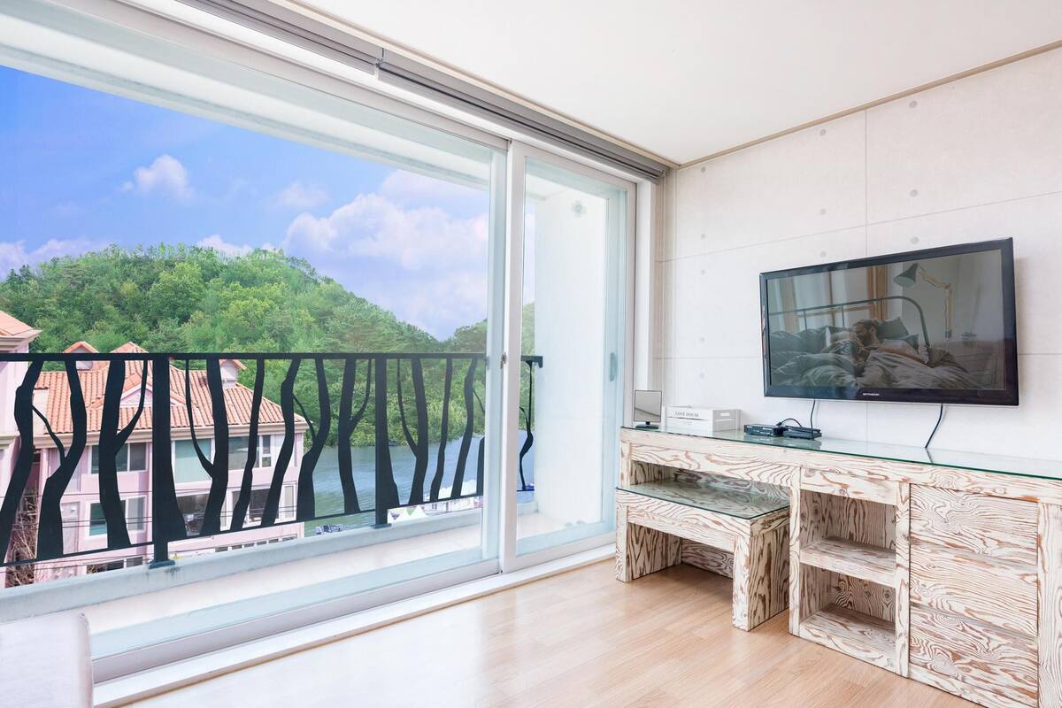 Een moderne woonkamer met een grote schuifpui naar een balkon, een flatscreen televisie aan de wand en een glazen salontafel.