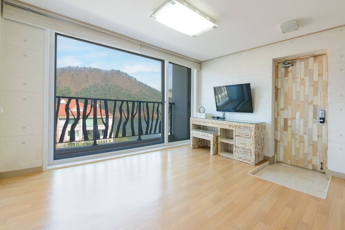 Een ruime kamer met een grote glazen schuifdeur naar een balkon, een houten bureau met een televisie, en een witte deur met een houten paneeldecoratie.