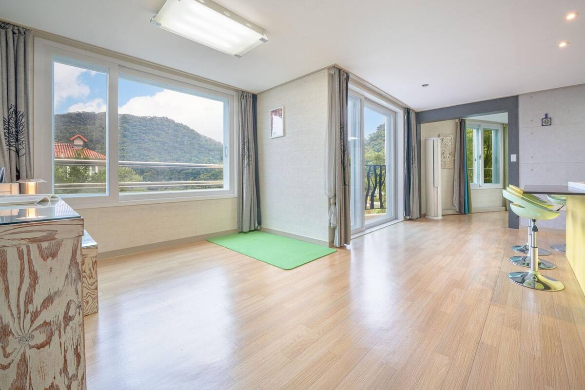Ruime kamer met grote ramen, een groene mat, een gele stoel, en een keuken met een geel kastje.