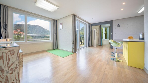 Ruime kamer met grote ramen, een groene mat, een gele stoel, en een keuken met een geel kastje.