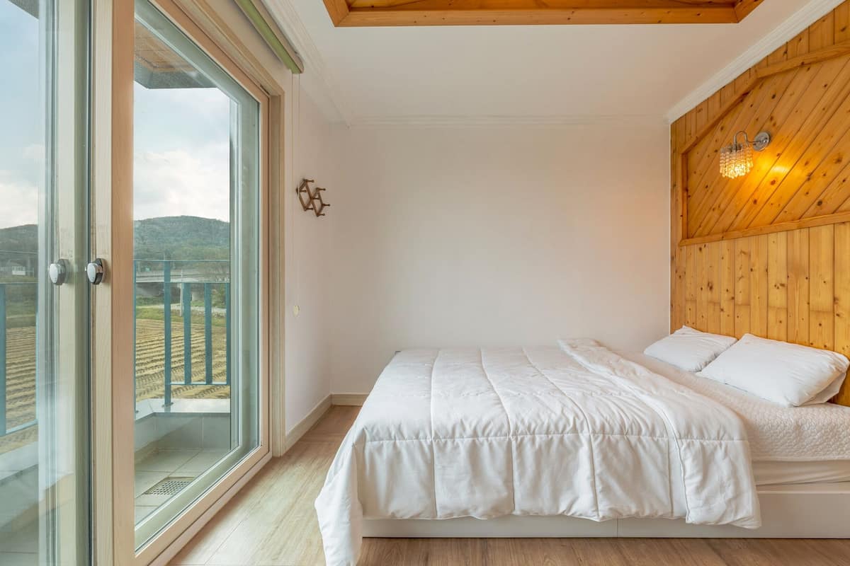 A bedroom with a large window, wooden ceiling, and a bed with white bedding.