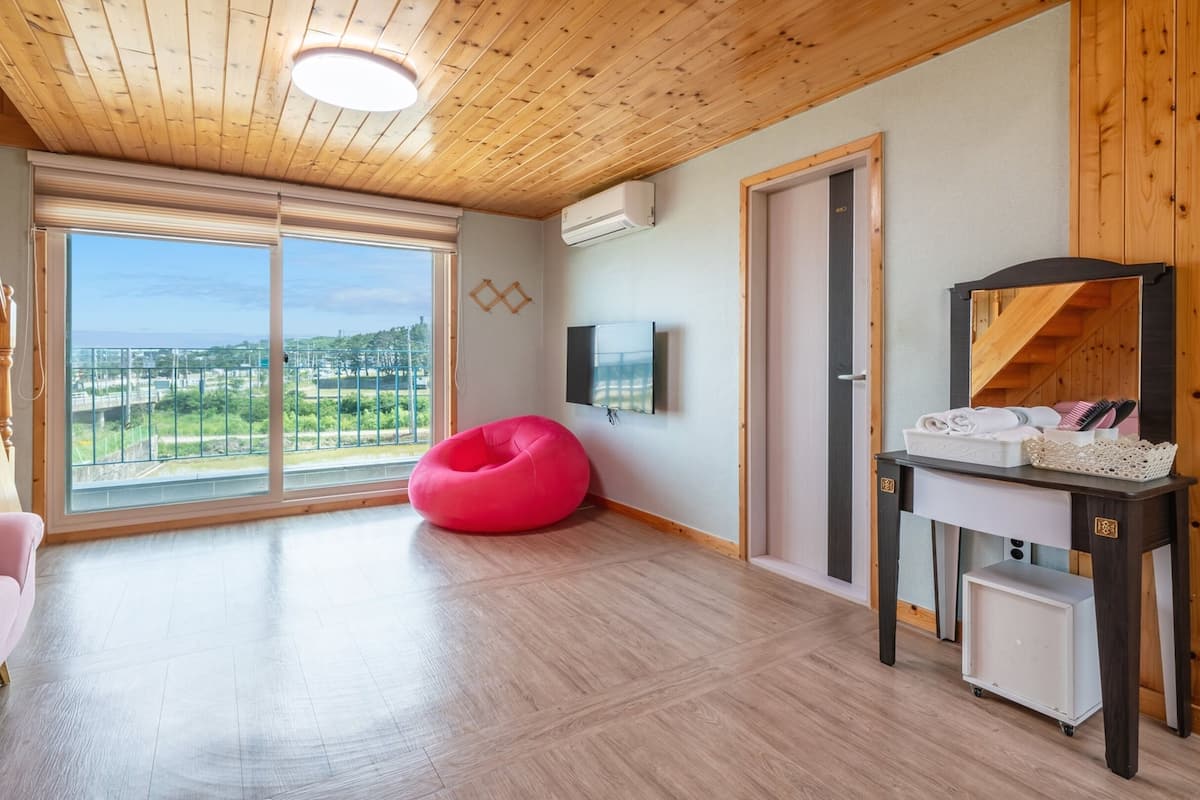 A spacious room with a large window, a pink bean bag, a wooden ceiling, and a small table with towels.