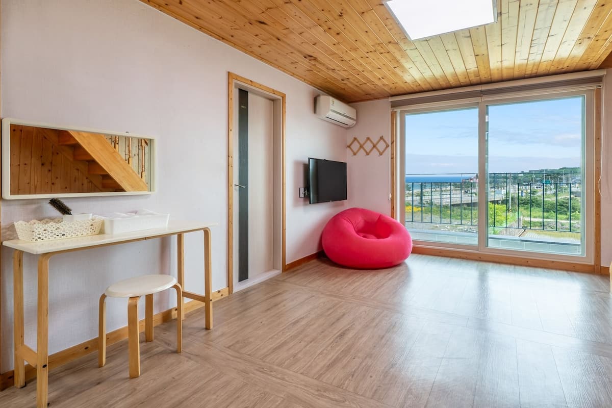 A modern living room with a wooden floor, a red bean bag chair, a small white table with a basket, a TV mounted on the wall, and a large window with a view of the outdoors.