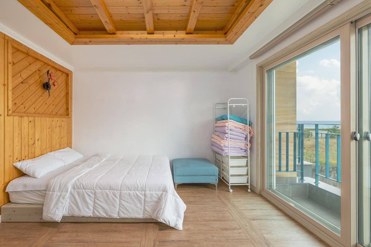 A bedroom with a wooden ceiling, a bed with white bedding, a blue bench, and a rack of folded clothes.