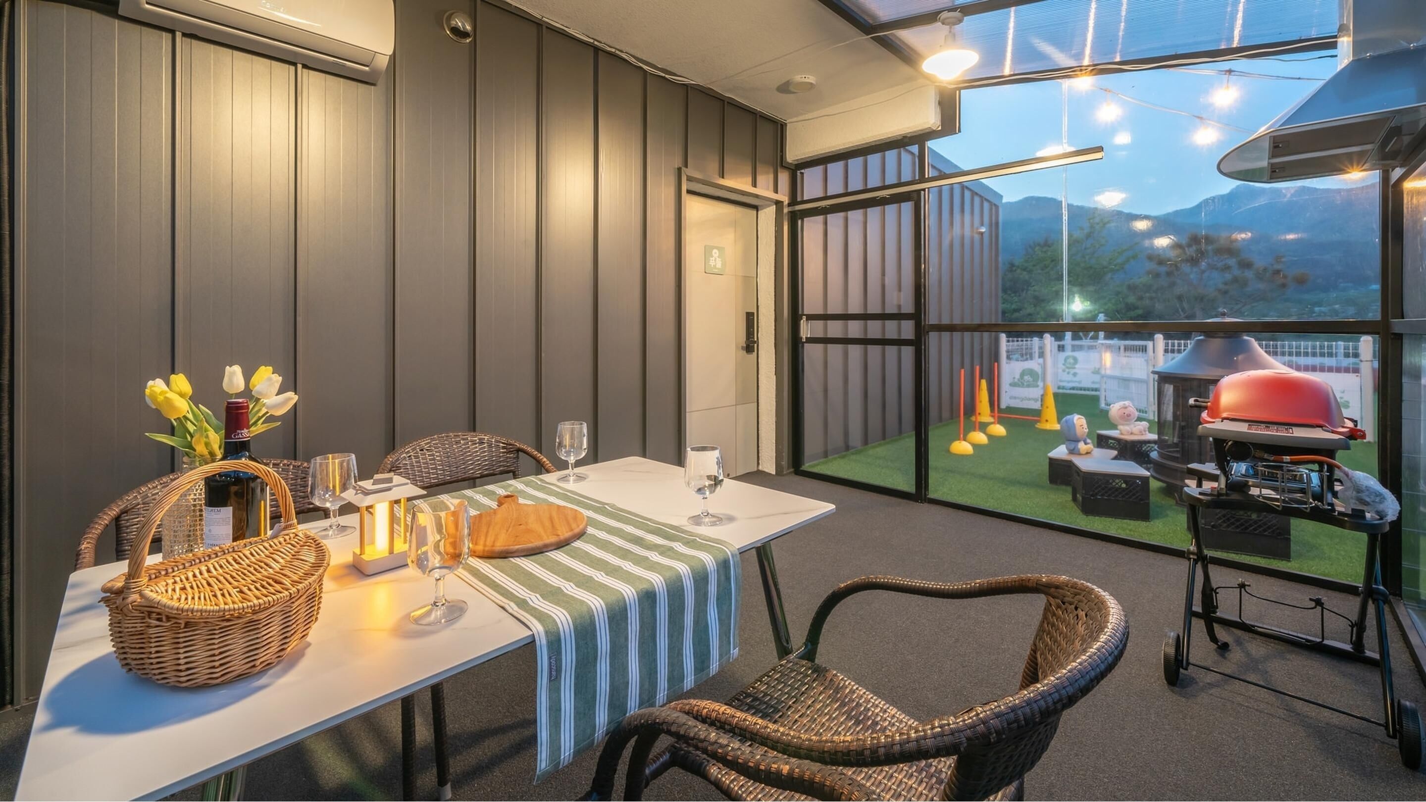 Un coin repas avec une table dressée pour deux personnes, agrémenté de paniers en osier, de verres à vin et d’une bouteille de vin. Une grande fenêtre offre une vue sur un espace barbecue extérieur.