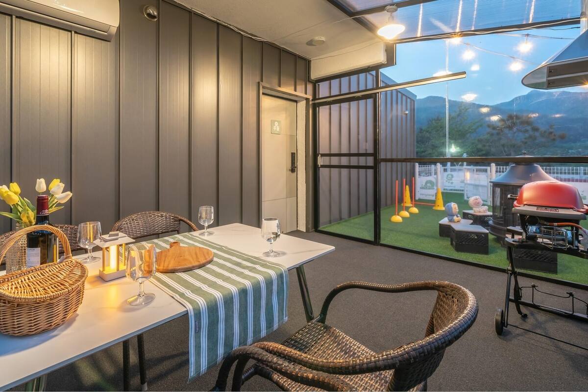 Un coin repas avec une table dressée pour deux personnes, agrémenté de paniers en osier, de verres à vin et d’une bouteille de vin. Une grande fenêtre offre une vue sur un espace barbecue extérieur.