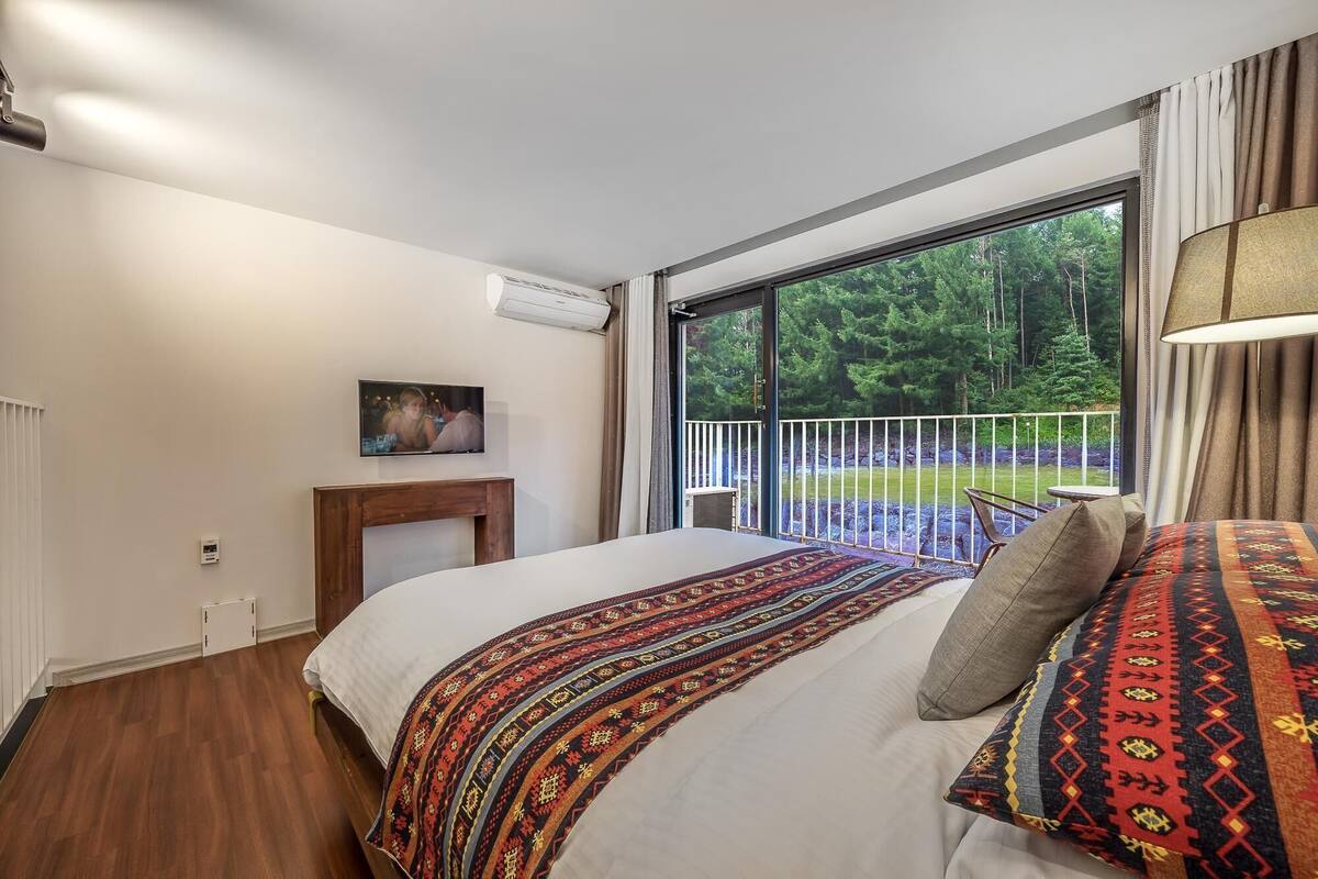 Une chambre avec une grande fenêtre, un lit, une table de chevet et une vue sur une forêt.