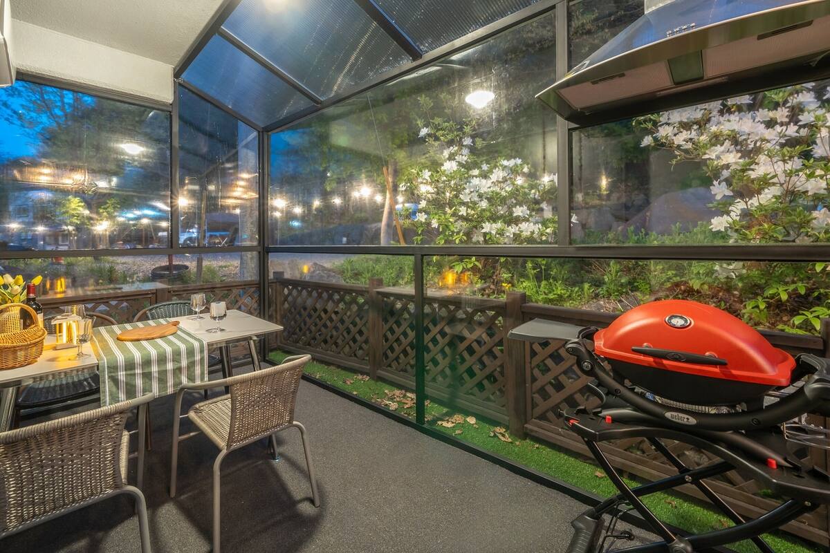 Un espace extérieur aménagé pour prendre les repas, avec une table pour deux, des chaises en osier, un barbecue et une vue sur la rue et les arbres, grâce à de grandes fenêtres.